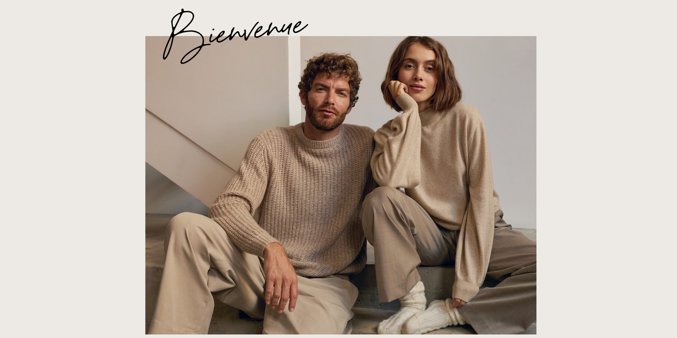 Deux modèles assis, vêtus de pulls en maille beige et de pantalons neutres, dans un décor minimaliste.