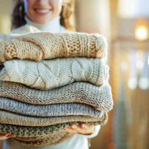 Closeup on happy elegant middle age housewife in white sweater and skirt at modern home in sunny winter day holding pile of sweaters.